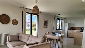 Uma sala de estar moderna com um sofá bege, mesa de jantar e cadeiras. Há uma área de cozinha com geladeira e máquina de café.