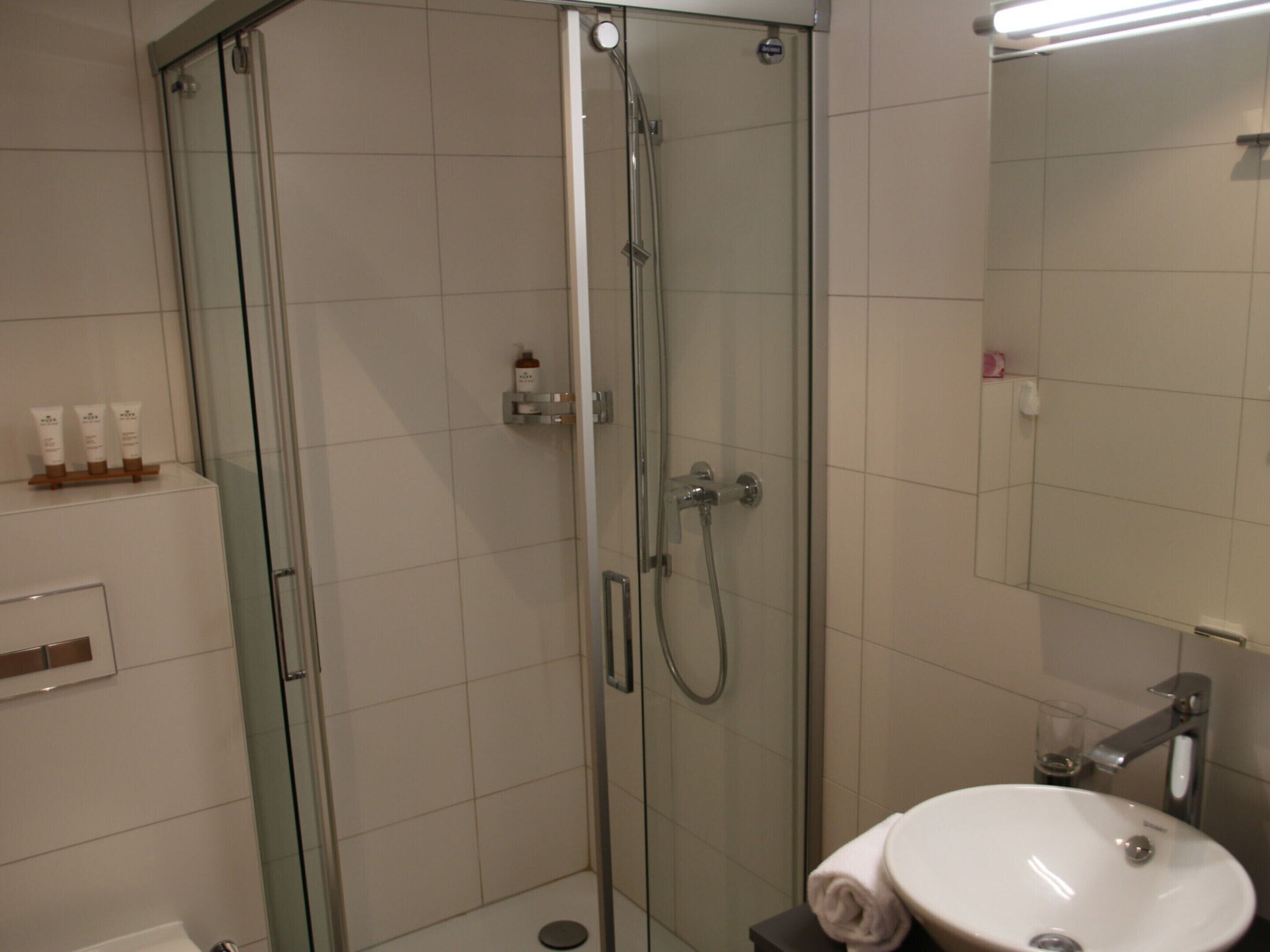 Ein modernes Badezimmer mit einer Duschkabine aus Glas, einem weißen Waschbecken und Pflegeprodukten auf einem Regal.