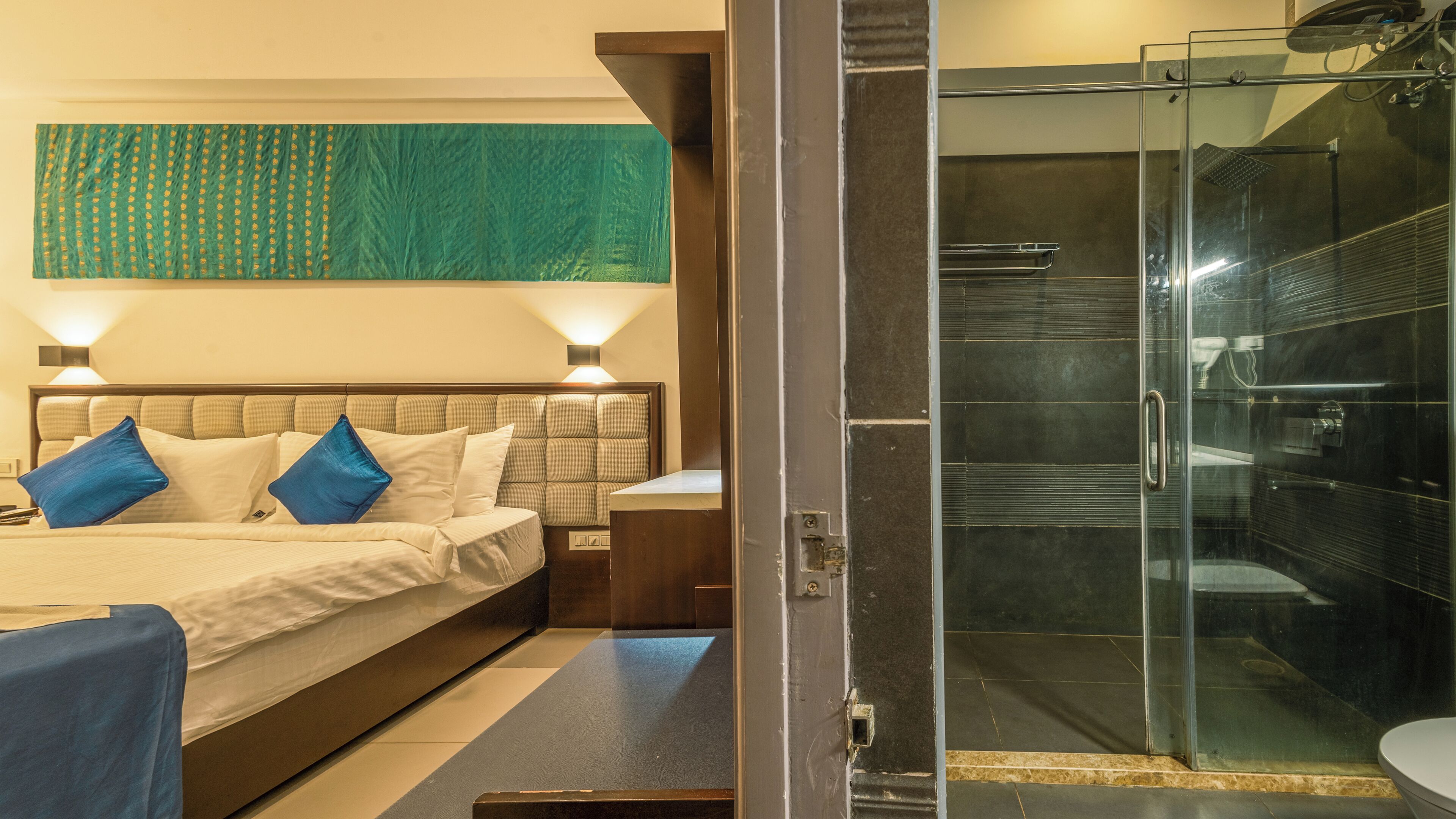 A hotel room with a bed, bedside lamps, a large green abstract painting, and a glass-enclosed shower area.