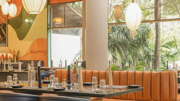 A restaurant with a long table set for dining, cushioned seating, and large windows with a view of greenery.