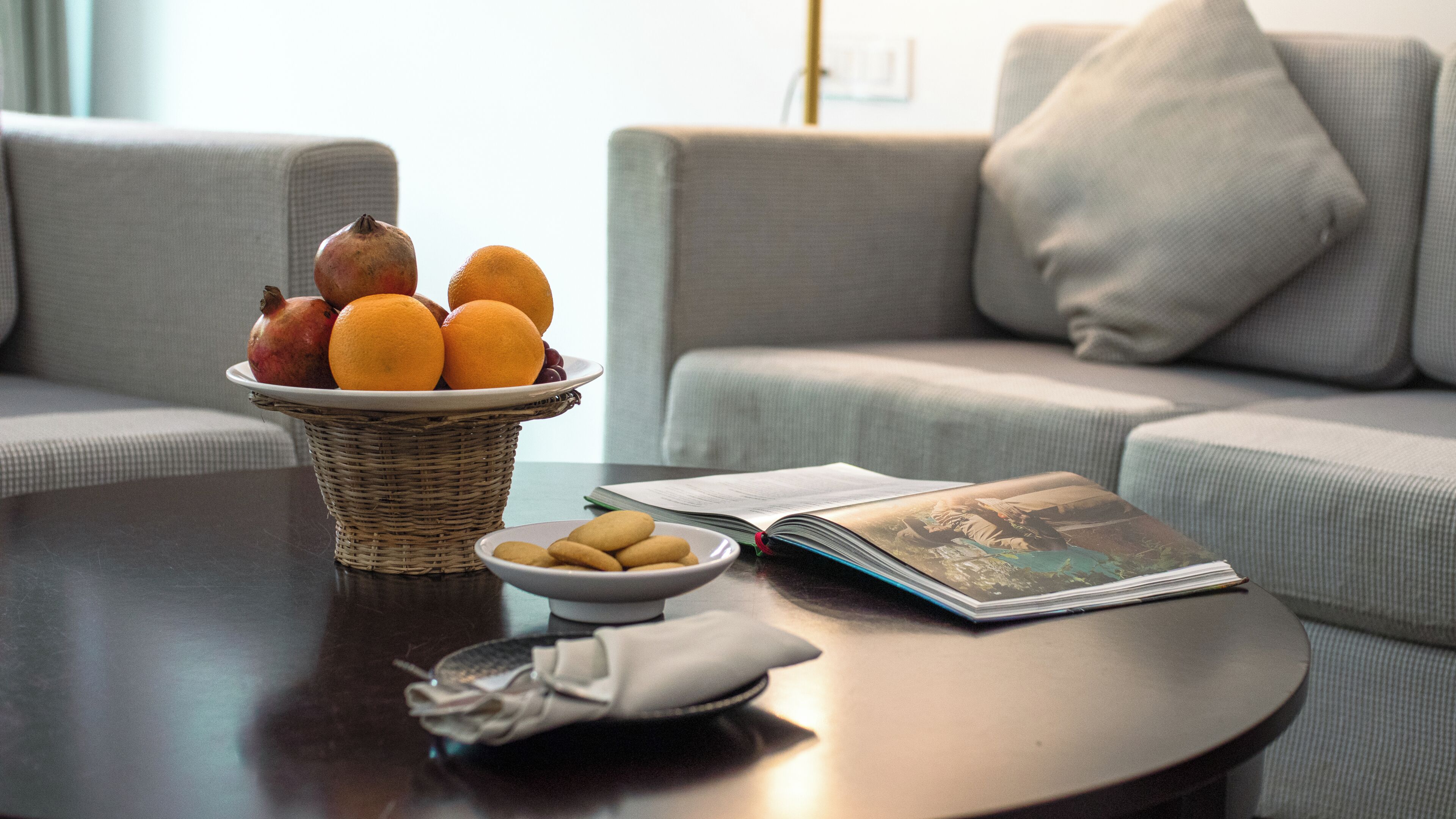 A living room with a sofa, a coffee table with a basket of fruit, a bowl of cookies, and an open magazine.