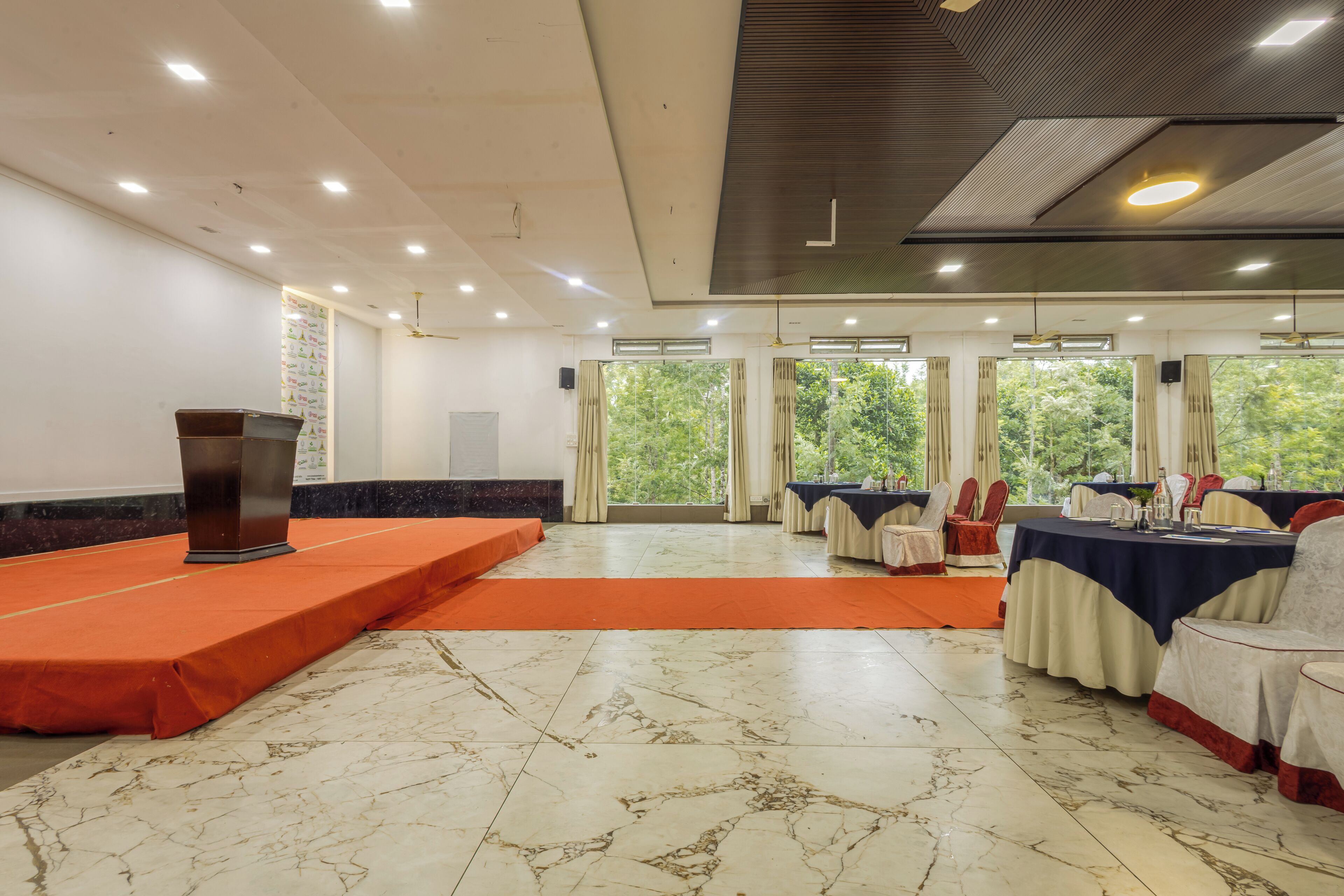 Un salón de conferencias con un atril, mesas con sillas y amplias ventanas con cortinas.