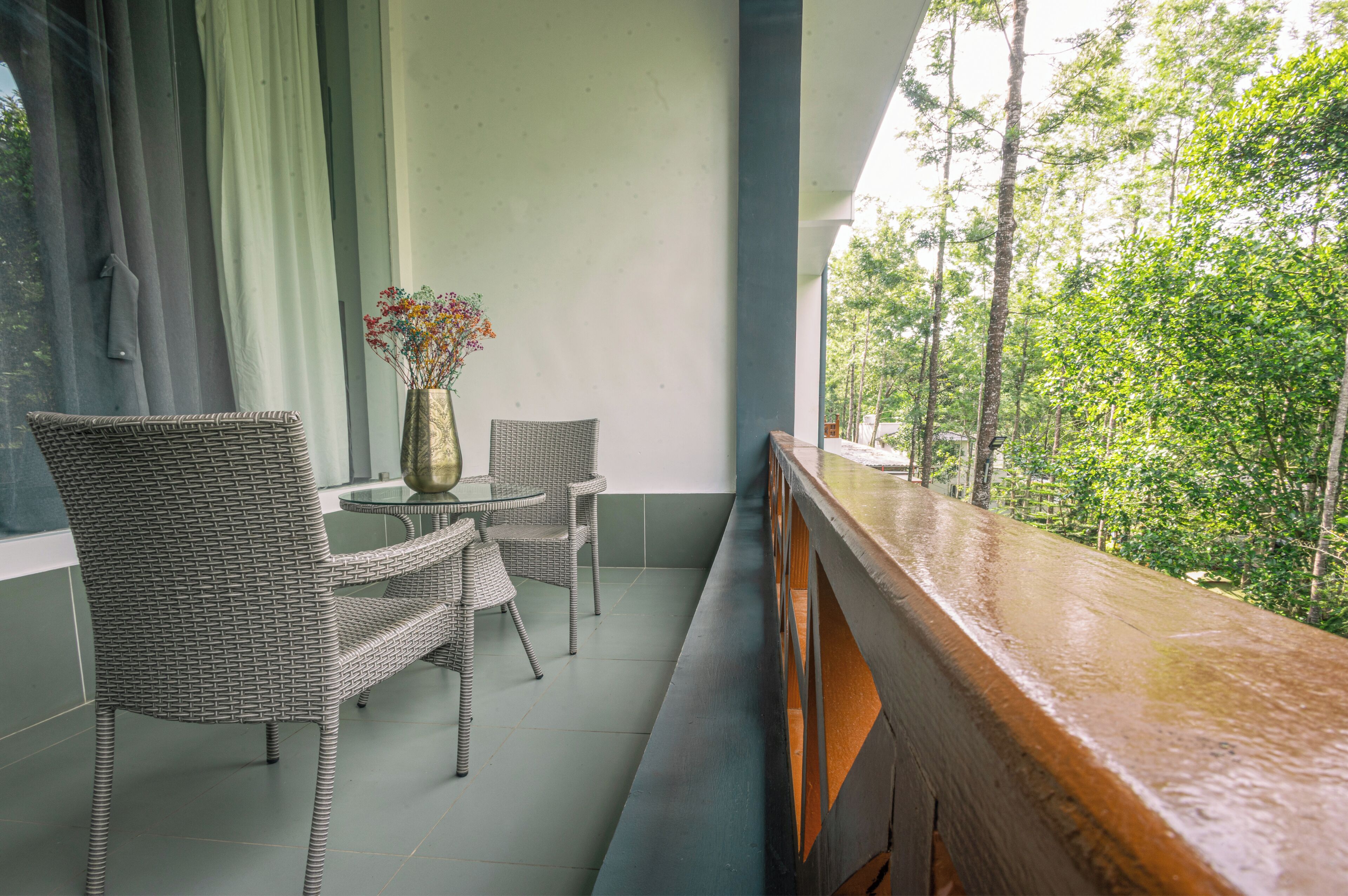 Een balkon met rieten stoelen, een tafeltje, een bloevas en uitzicht op bomen.
