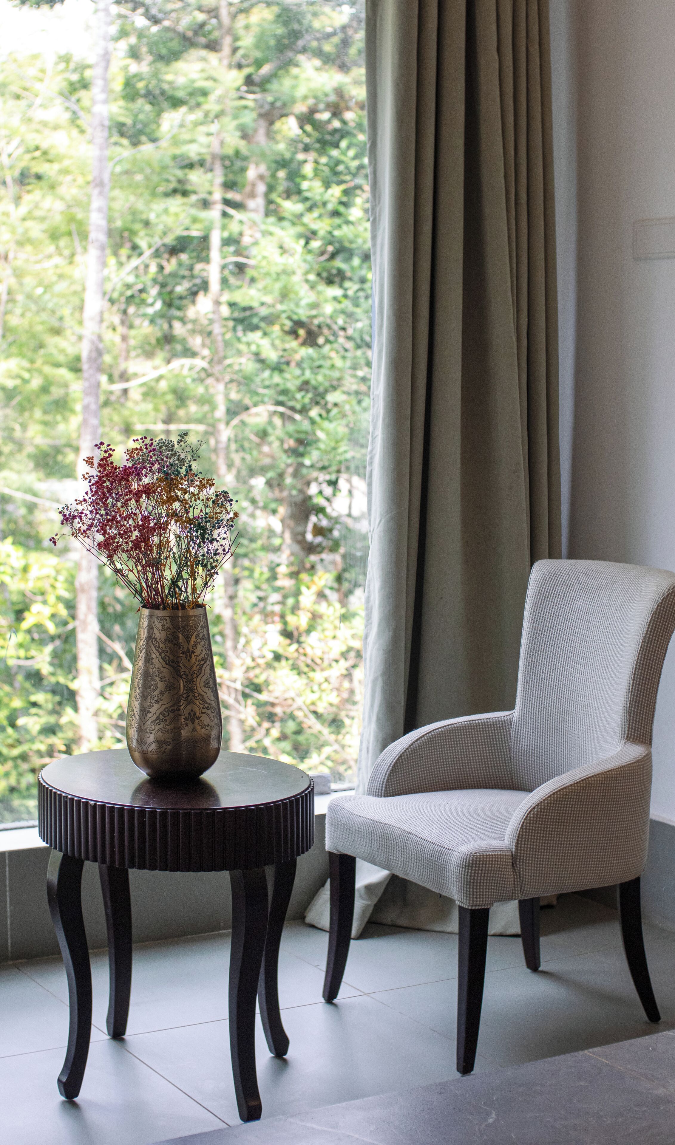 Ein Zimmer mit Blick auf Bäume, ein Sessel und ein Tisch mit Blumen in einer Vase.