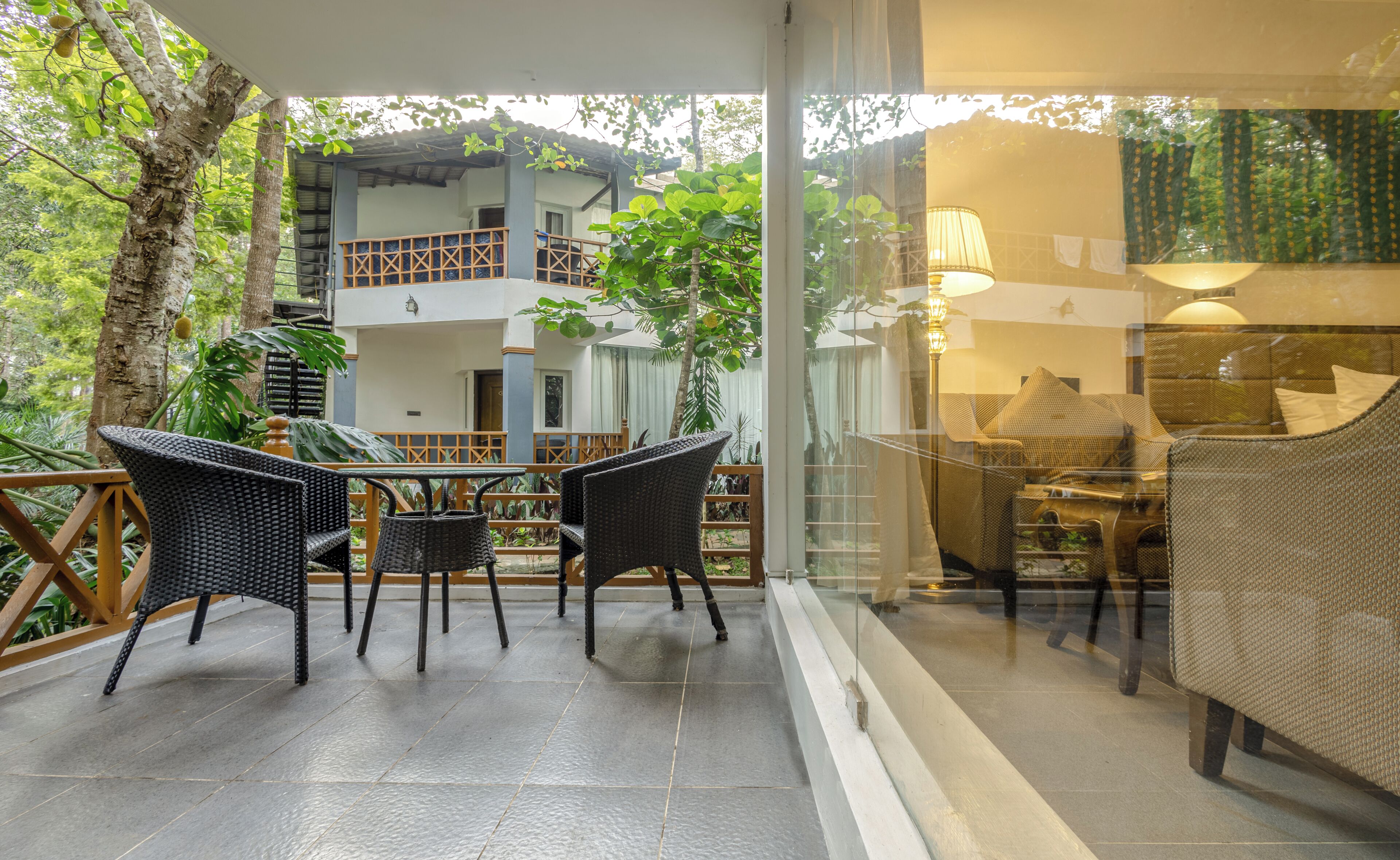 A covered outdoor seating area with wicker furniture, a glass partition, and a view of a building with a balcony.