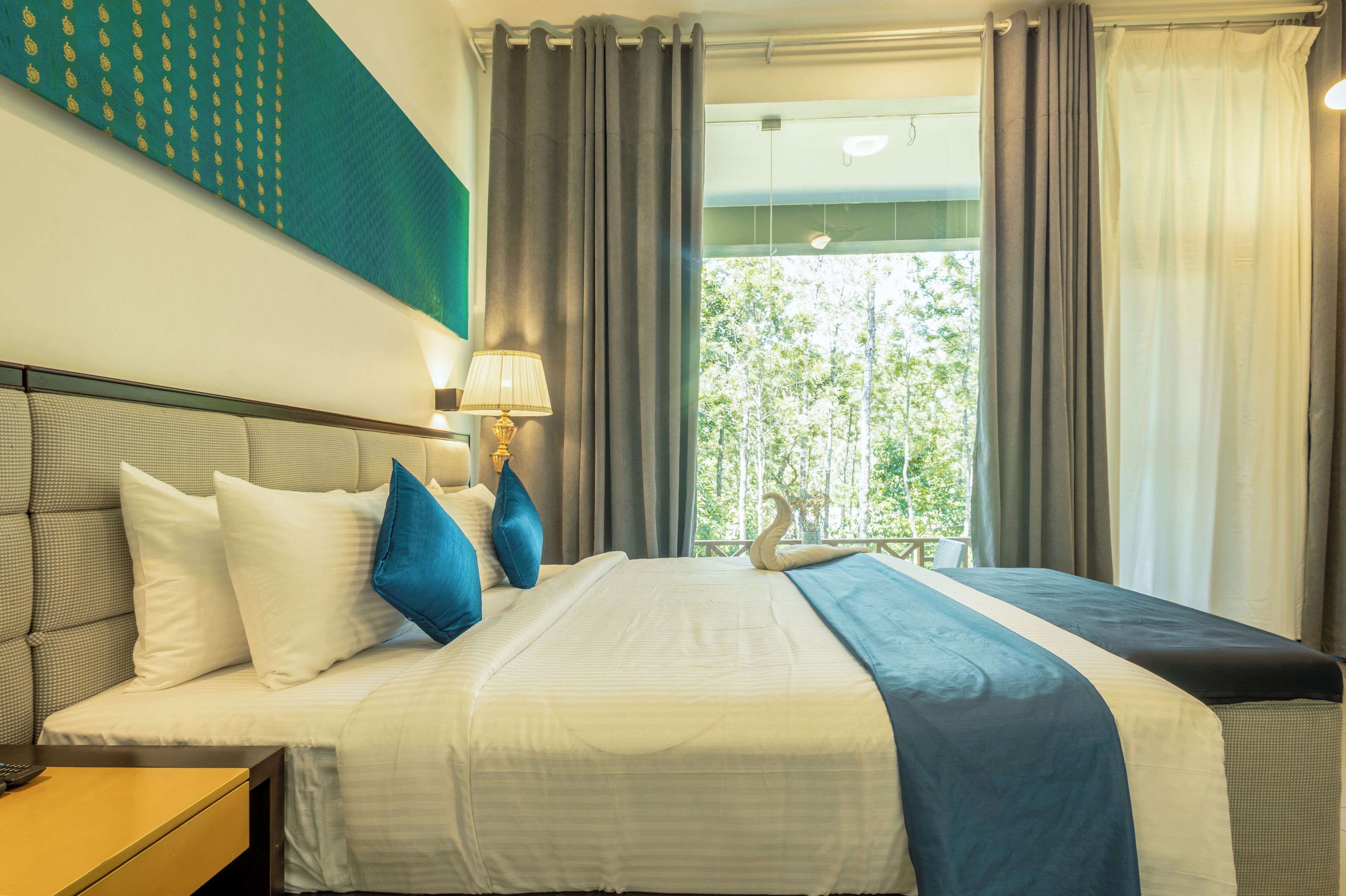 A hotel room with a bed, a nightstand, a lamp, and a view of trees through the window.