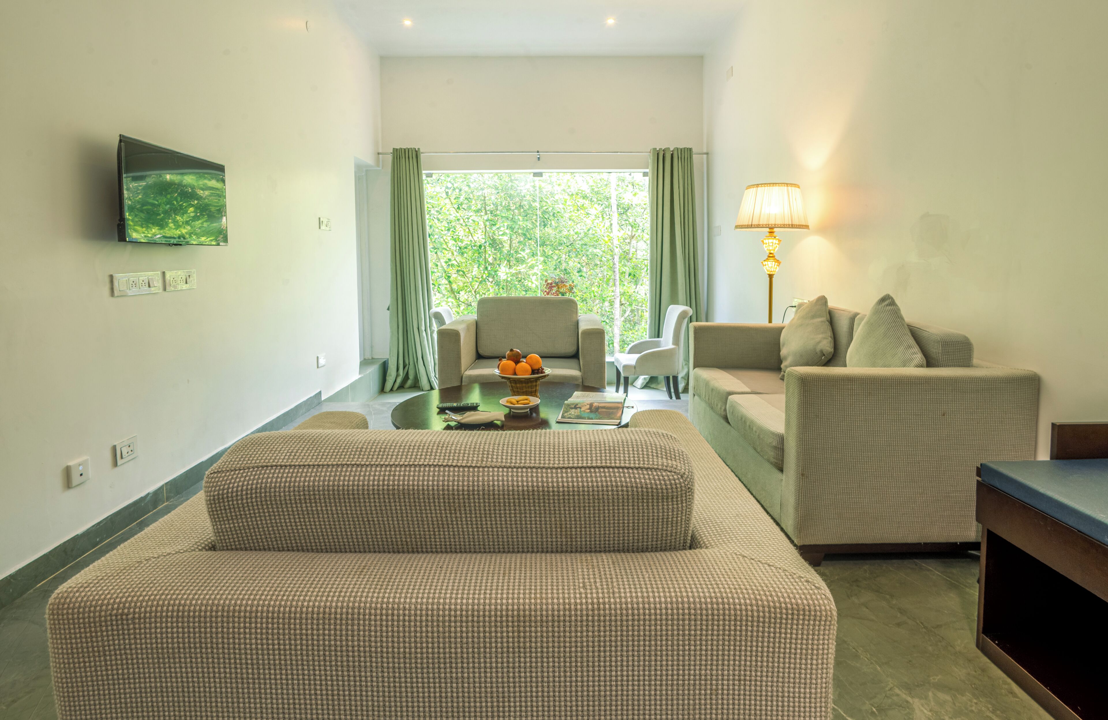 A living room with a sofa, armchairs, a coffee table, a TV mounted on the wall, and a window with curtains.