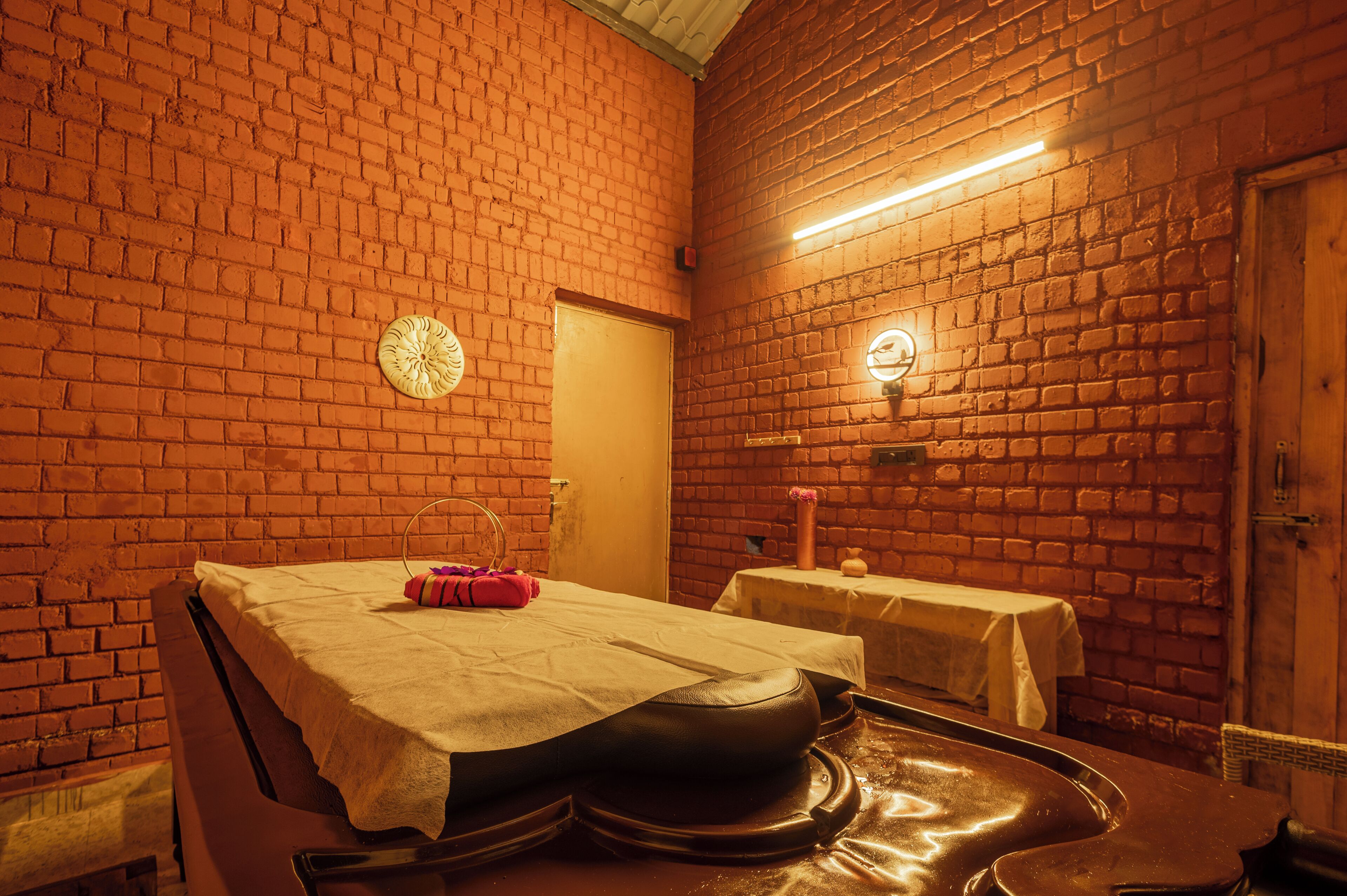 A room with a brick wall, a massage table, a small table with a tablecloth, and a wooden door.