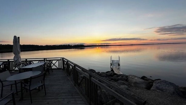 Eine Holzterrasse mit Tischen und Stühlen, die auf ruhiges Gewässer im Sonnenuntergang blickt.