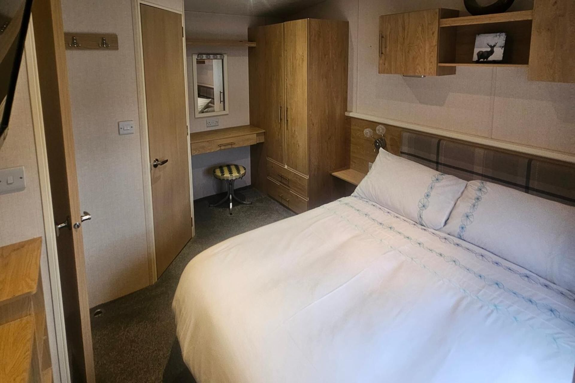 A bedroom with a bed, a wooden wardrobe, a small stool, a mirror, and a shelf with a picture.