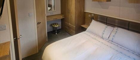 A bedroom with a bed, a wooden wardrobe, a small stool, a mirror, and a shelf with a picture.