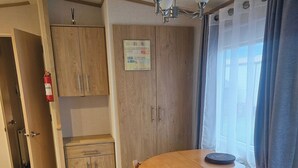 A compact room with a wooden dining table, chairs, a small cabinet, a ceiling light, and a window with curtains. - Dunooncaravan/2bed/sleeps4/scenicwwalks/tv/wifi (Dunoon)