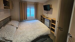 A bedroom with a bed, a television mounted on the wall, a window with curtains, and a door. - Dunooncaravan/2bed/sleeps4/scenicwwalks/tv/wifi (Dunoon)