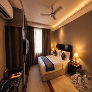 Quarto de hotel com cama, luminárias de cabeceira, ventoinha no teto, televisão e uma mesinha com um vaso e garrafas de água.