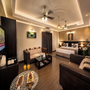 A modern hotel room with a sofa, a bed, a coffee table with snacks, a TV, and a ceiling fan. - Hotel The Grand Shoba (New Delhi)