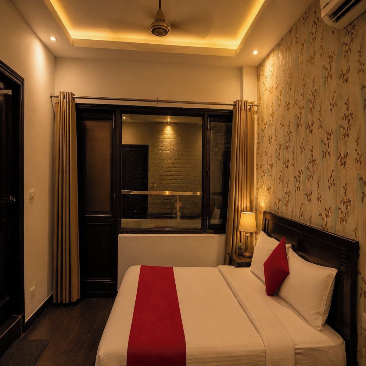 A hotel room with a bed, a window with curtains, a wall with floral wallpaper, and a ceiling light.