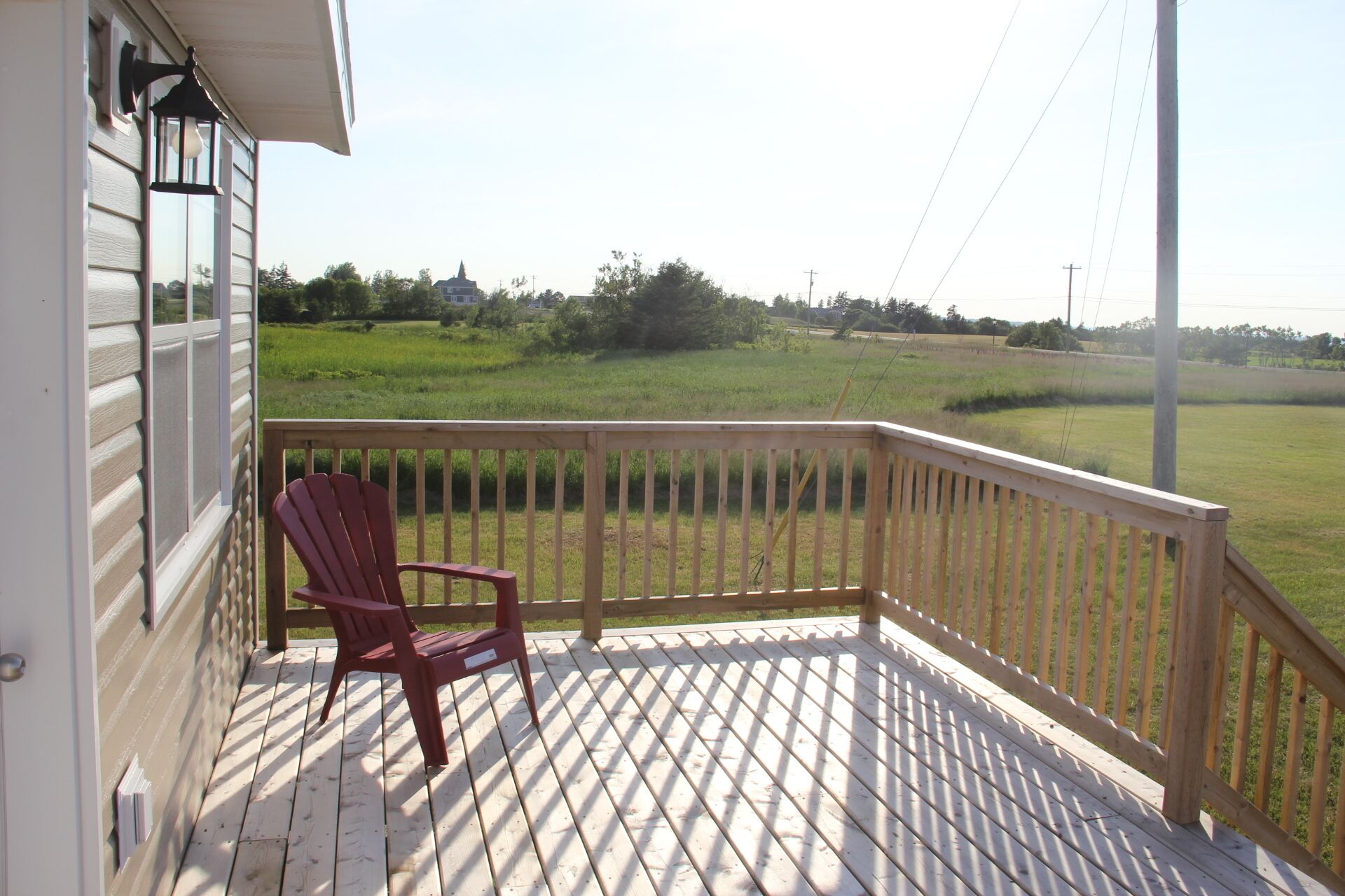 A wooden deck with a red Adirondack chair, a white railing, and a view of a green field.