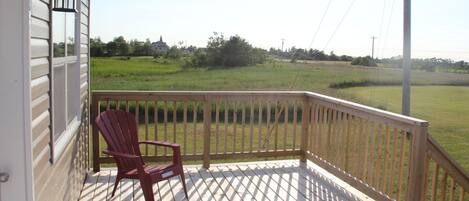 A wooden deck with a red Adirondack chair, a white railing, and a view of a green field.