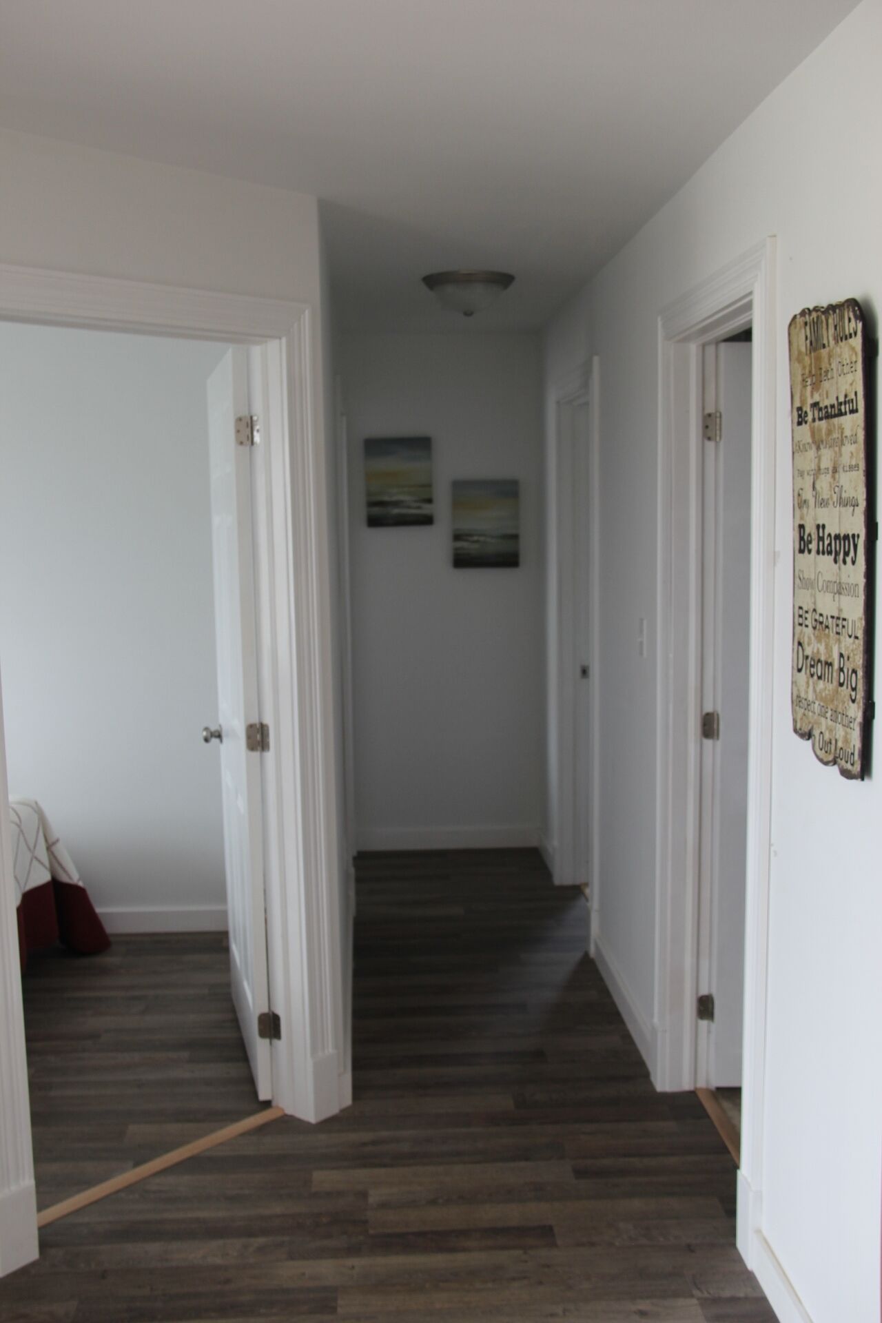 A hallway with wooden flooring, white walls, and two doors. There are two framed pictures on the wall and a sign with motivational phrases.