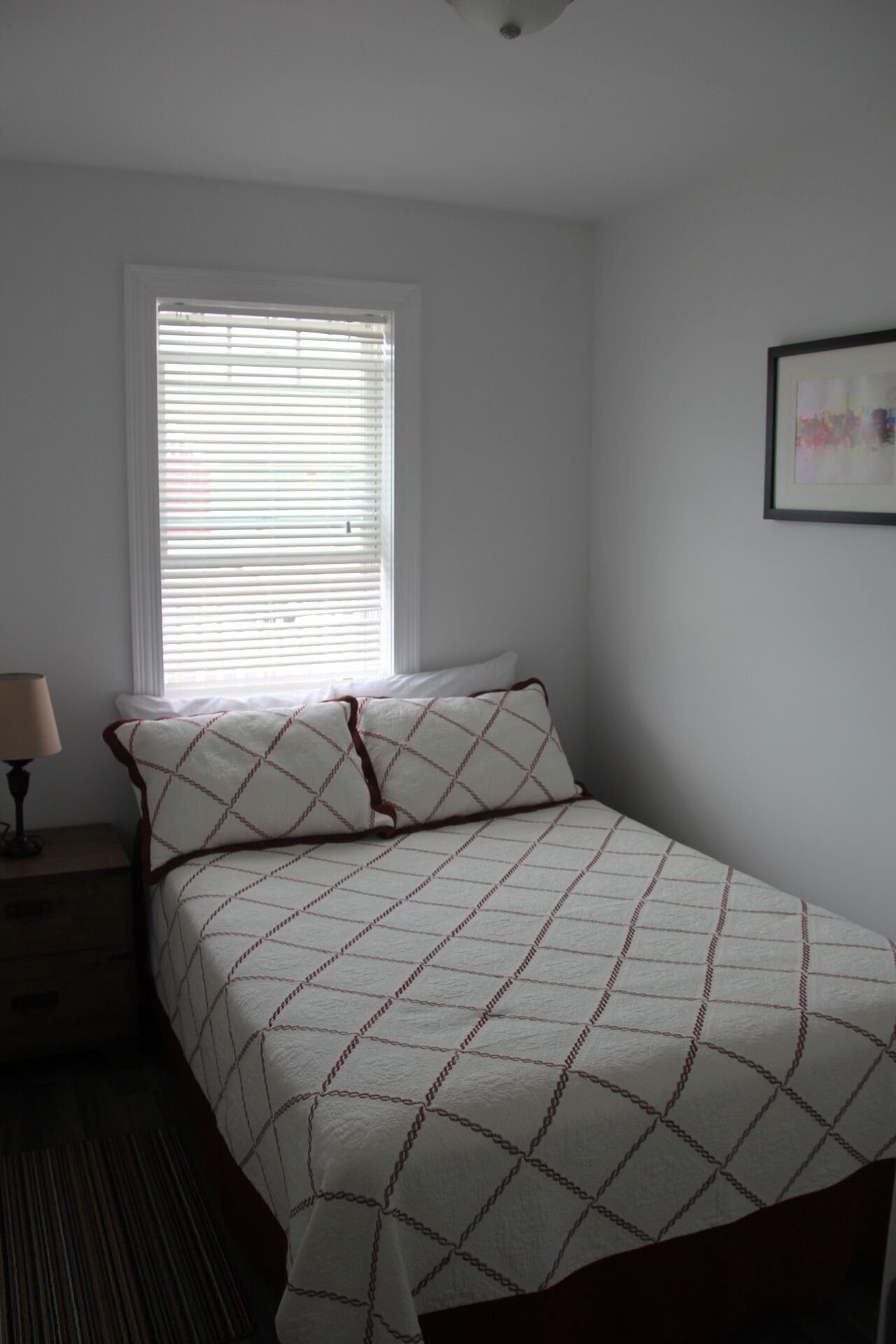 A bedroom with a bed, a nightstand, a lamp, a window with blinds, and a framed picture on the wall.