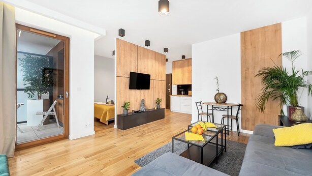 Una sala moderna con un sofá gris, una mesa baja con fruta, una mesa de comedor de madera y una zona de cocina con un televisor.