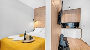 Una habitación de hotel moderna con una cama, una mesita con una botella y vasos, y una kitchenette con armarios y un televisor.