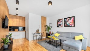 Un espacio habitable moderno con un sofá gris, suelo de madera, una zona de comedor con mesa y sillas, y una cocina con armarios de madera y un televisor de pantalla plana.