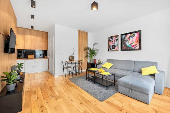 A modern living space with a grey sofa, wooden flooring, a dining area with a table and chairs, and a kitchen with wooden cabinets and a flat-screen TV. - Lumina stylish apart with balcony (Warsaw)
