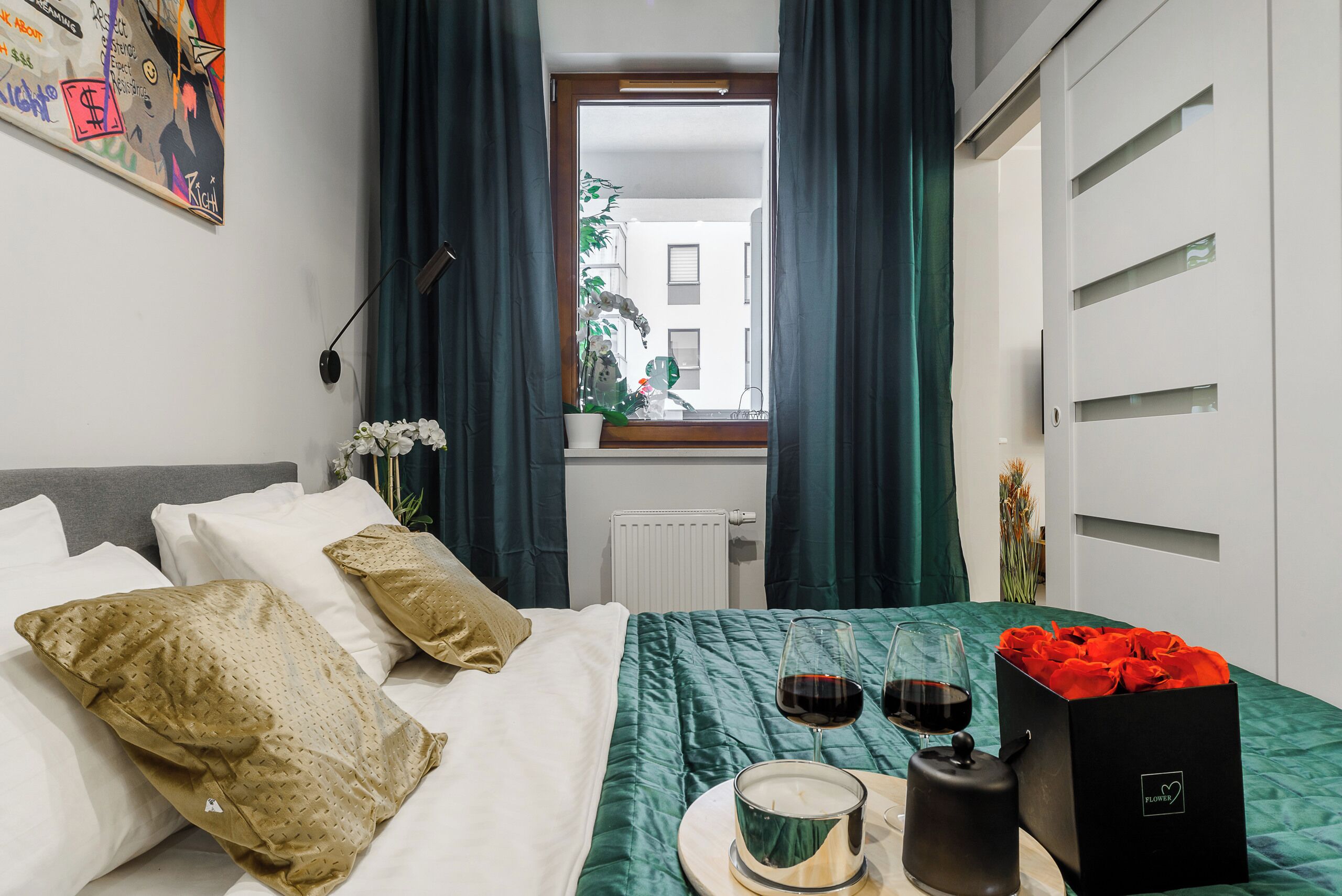 Et hotelværelse med en seng, et vindue med gardiner, et natbord med et stearinlys, et glas vin og en æske roser.