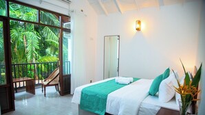 A bedroom with a bed, a nightstand, a mirror, a window with a view of greenery, and a balcony with a chair and table. - Aaradhya Madu River  (Ambalangoda)