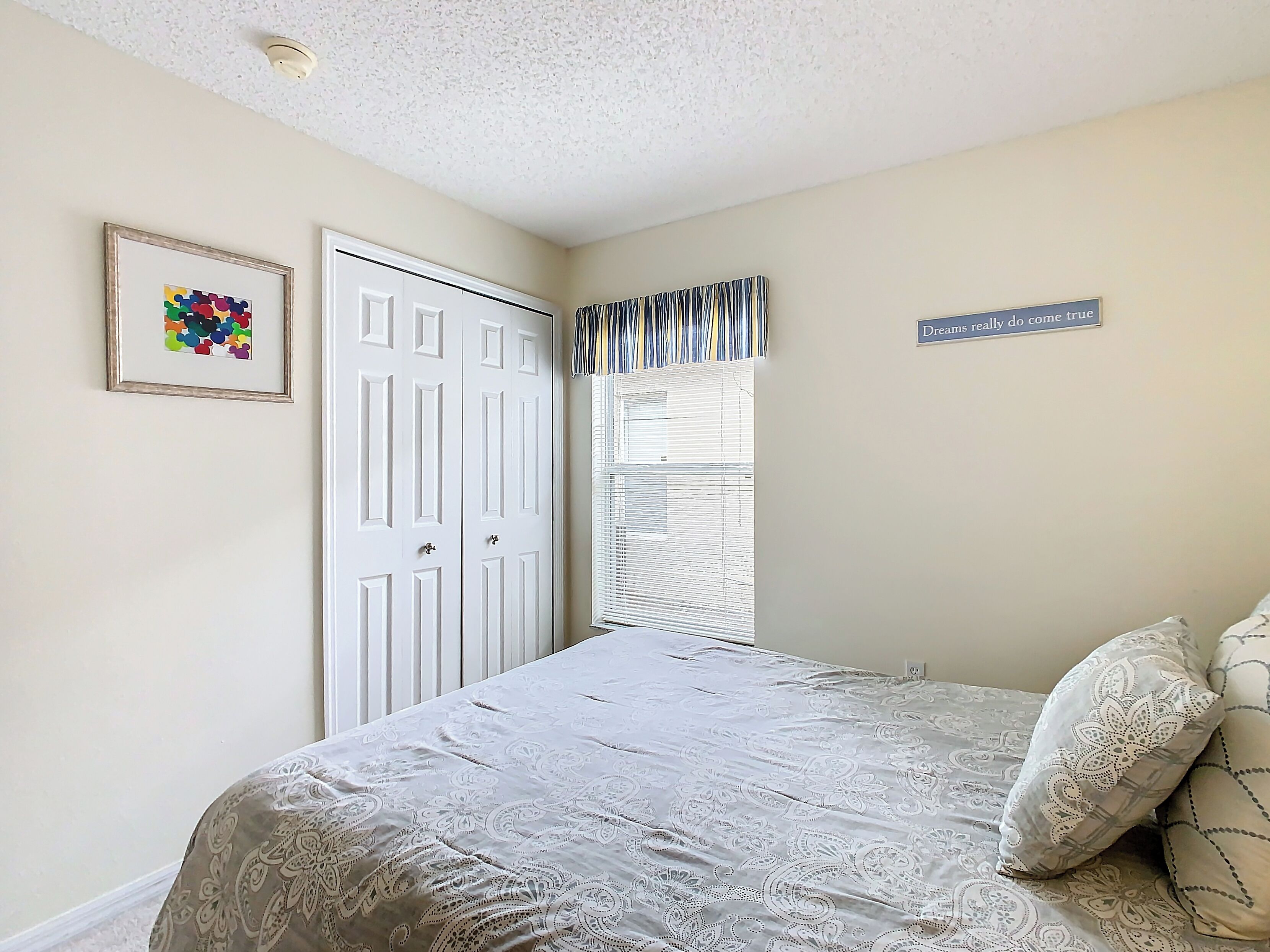 Een slaapkamer met een bed, een deur, een raam met zonwering, een wandbord en een plafondlamp.