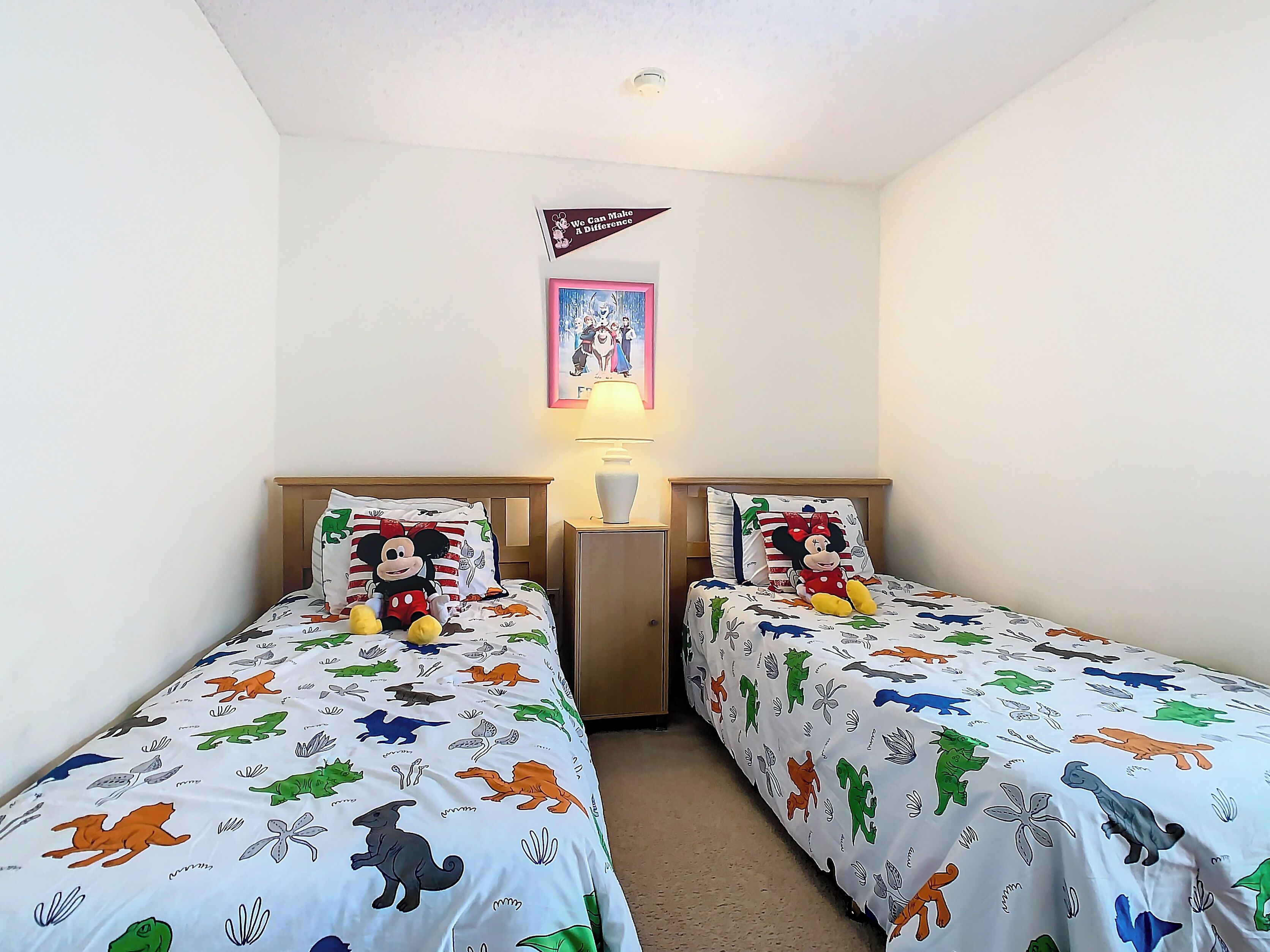 Une chambre avec deux lits, chacun agrémenté d’une couvre-lit et de coussins aux motifs de Mickey Mouse. Une petite table avec une lampe se trouve entre les lits, et un tableau encadré est accroché au-dessus de la table.