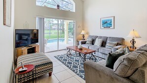 A spacious living room with a large TV, comfortable seating, a coffee table, and a view of a backyard through large windows. - Golfview 4 bedroom villa with pool in Southern Dunes, 20 miles from Disneyworld (Haines City)