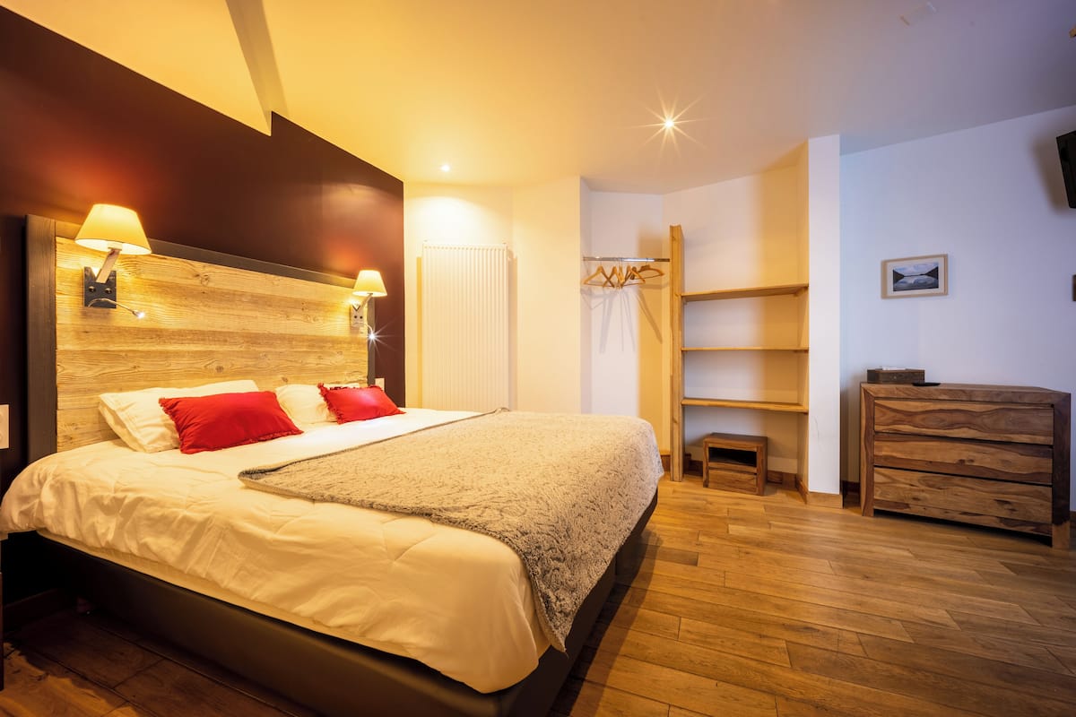 A bedroom with a bed, bedside lamps, a wooden headboard, a wooden dresser, a shelf, and a hanging garment rack.