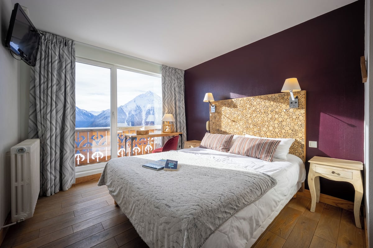 A bedroom with a large bed, two bedside lamps, a TV mounted on the wall, and a view of mountains through the window.