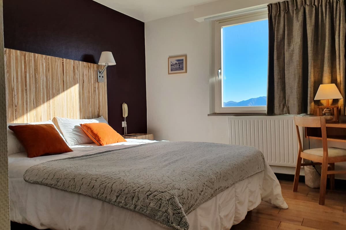 A hotel room with a bed, a desk, a chair, a window with curtains, a lamp, and a picture on the wall.