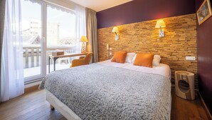 A hotel room with a large bed, two bedside lamps, a wooden accent wall, a desk with a chair, and a view through a large window. - Hôtel Le Christiania Saint Lary Soulan (Saint-Lary-Soulan)