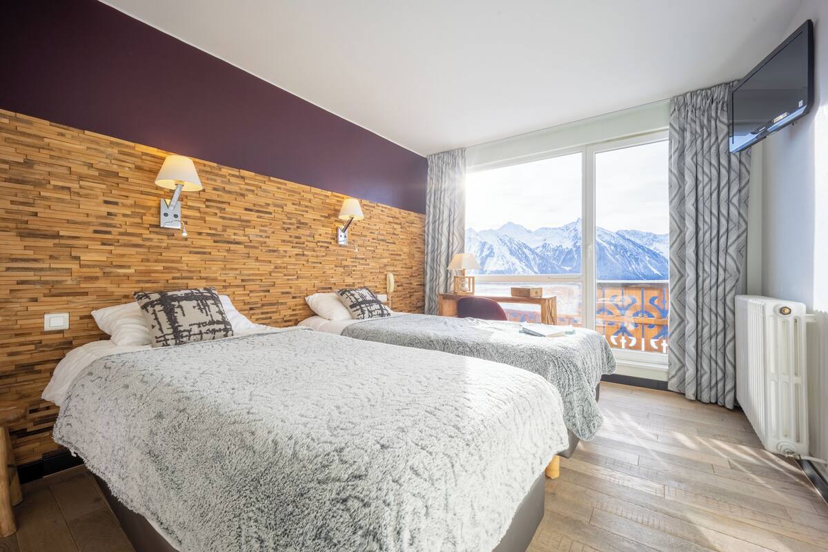A hotel room with two beds, a wooden wall, a TV, and a view of mountains.