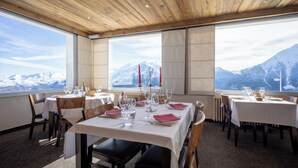 A dining area with tables set for a meal, large windows with a mountain view, and wooden ceiling beams. - Hôtel Le Christiania Saint Lary Soulan (Saint-Lary-Soulan)