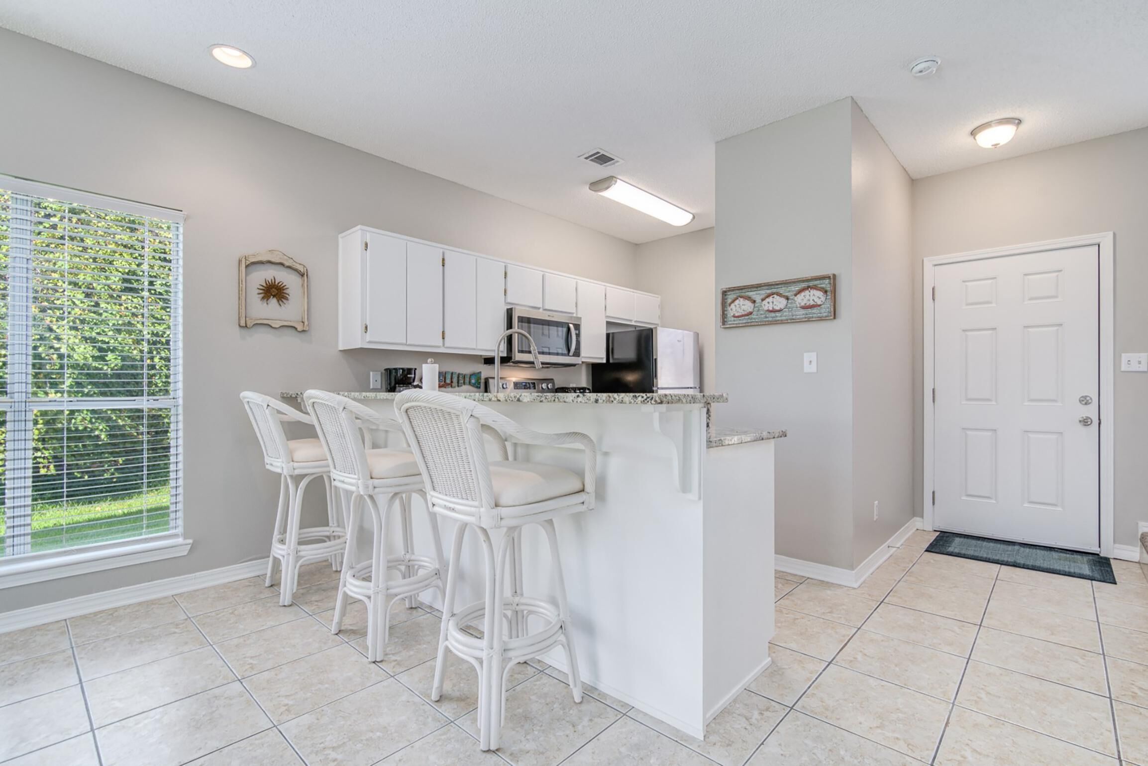 A kitchen with white cabinets, a microwave, and a refrigerator. There are three white bar stools.