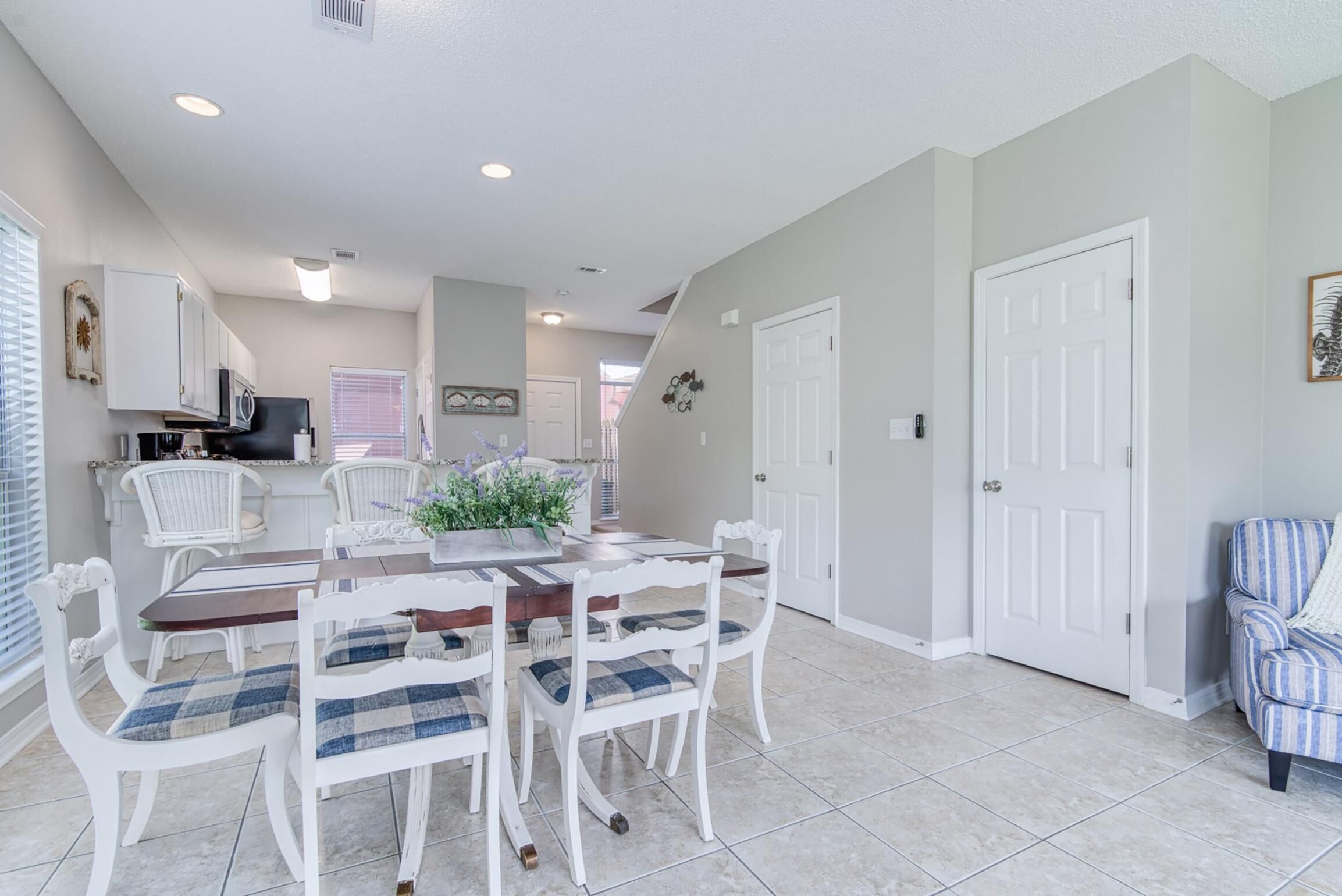 A dining area with a wooden table and white chairs, a kitchen in the background, and a door to the right.