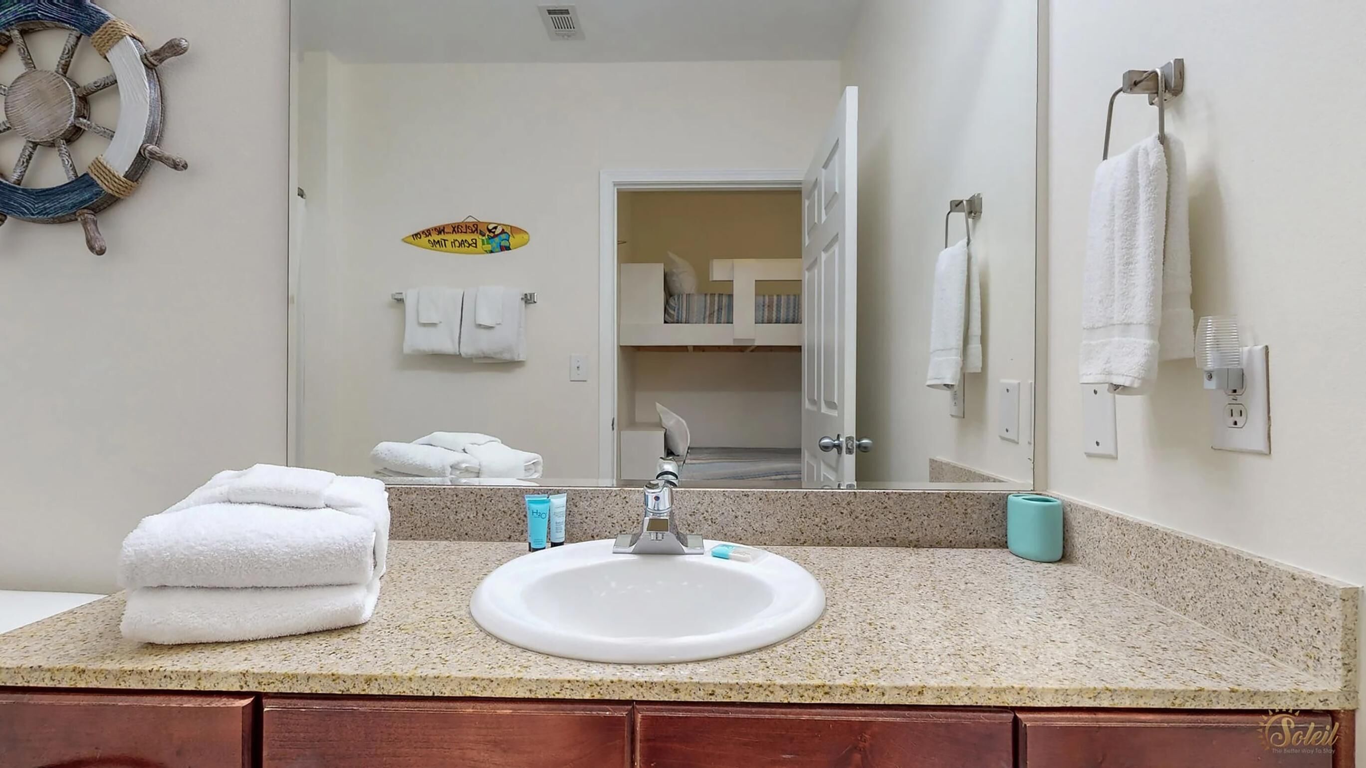 A bathroom with a granite countertop, white sink, and a mirror. There are towels stacked on the counter and a towel rack on the wall.