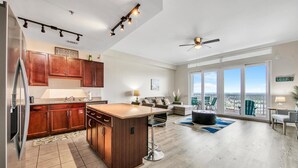A modern kitchen with wooden cabinets, a stainless steel refrigerator, and a large island. There is a dining area with a table and chairs, and a balcony with a view.