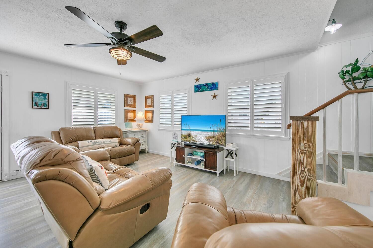 A spacious living room with leather sofas, a ceiling fan, a flat-screen TV, and a wooden staircase.