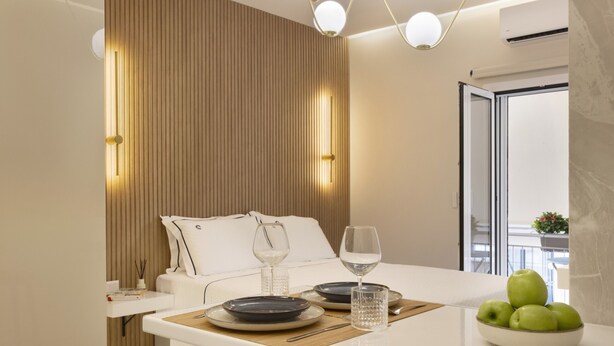 Quarto de hotel moderno com cama, mesa de cabeceira e área de refeições, incluindo uma tigela de maçãs e copos de vinho.