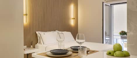Una habitación de hotel moderna con cama, mesita de noche y un área de comedor con un bol de manzanas y copas de vino.