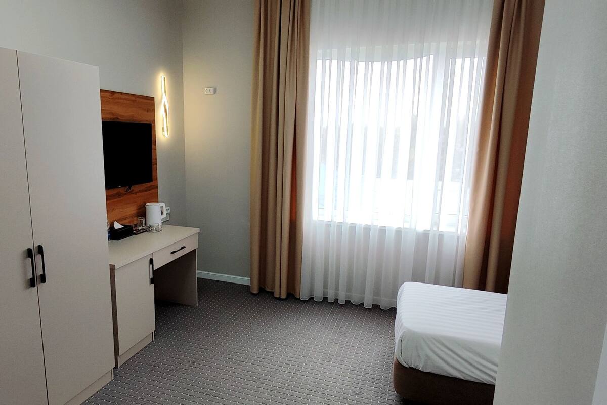 A hotel room with a wardrobe, a desk with a flat-screen TV, a bed, and a window with curtains.