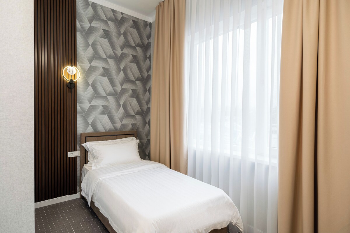 Habitación de hotel con una cama, una lámpara fijada a la pared, pared con diseño geométrico y cortinas traslúcidas.