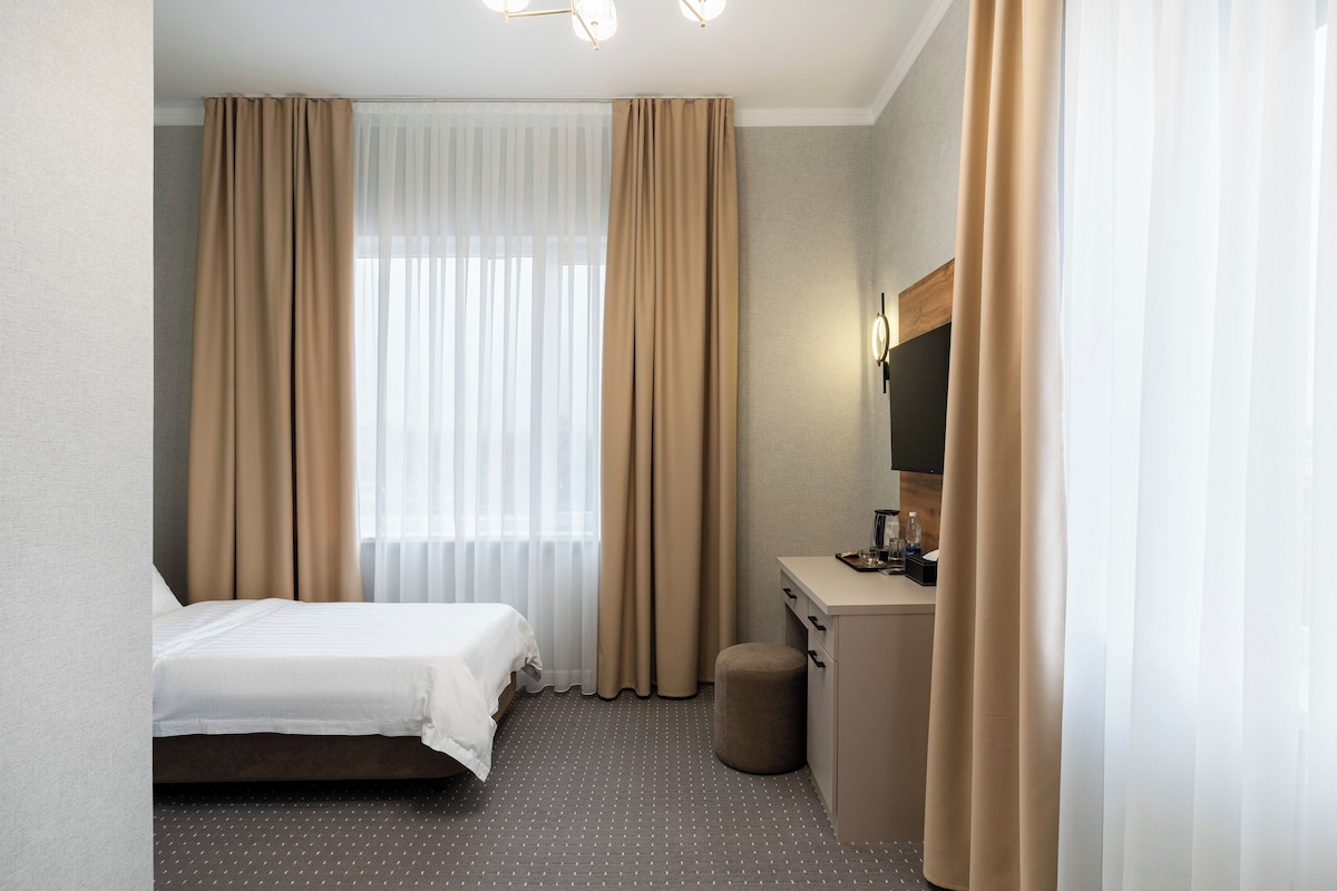 A hotel room with a bed, a desk, a chair, a small ottoman, and a mirror.