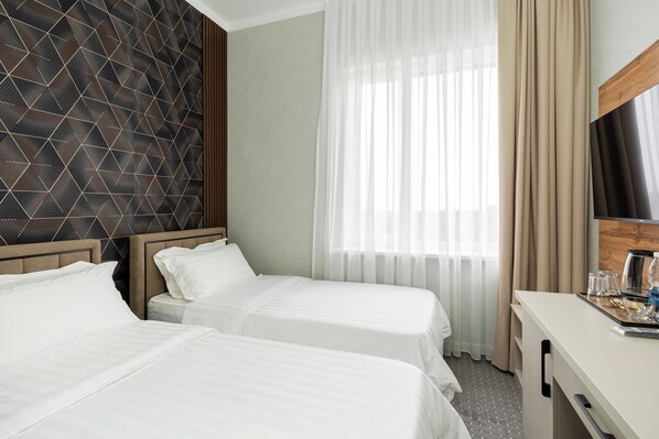 A hotel room with two beds, a wooden headboard, a large window with sheer curtains, a wooden desk, and a flat-screen TV.